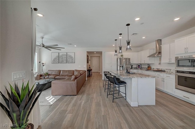 kitchen with an island with sink, white cabinets, hanging light fixtures, stainless steel appliances, and wall chimney exhaust hood