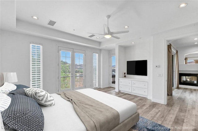 bedroom with light hardwood / wood-style flooring, access to outside, a multi sided fireplace, a tray ceiling, and ceiling fan