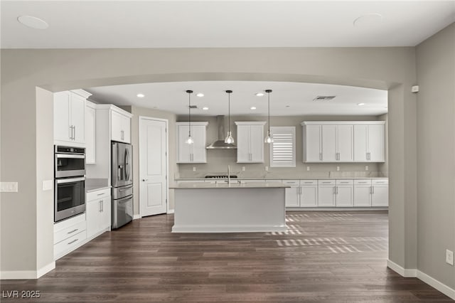 kitchen featuring pendant lighting, wall chimney range hood, white cabinetry, stainless steel appliances, and an island with sink