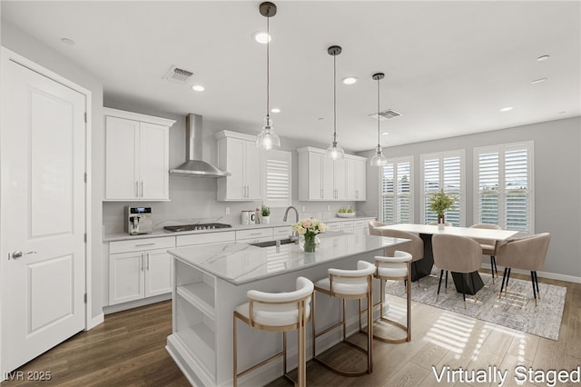 kitchen with sink, white cabinetry, decorative light fixtures, an island with sink, and wall chimney range hood