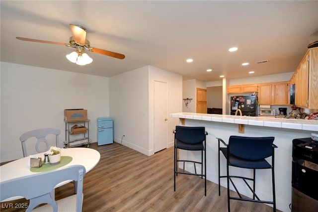 kitchen featuring a kitchen breakfast bar, tile counters, light hardwood / wood-style floors, black fridge, and light brown cabinets