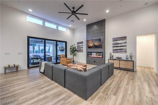 living room with ceiling fan, a towering ceiling, a tiled fireplace, and light wood-type flooring