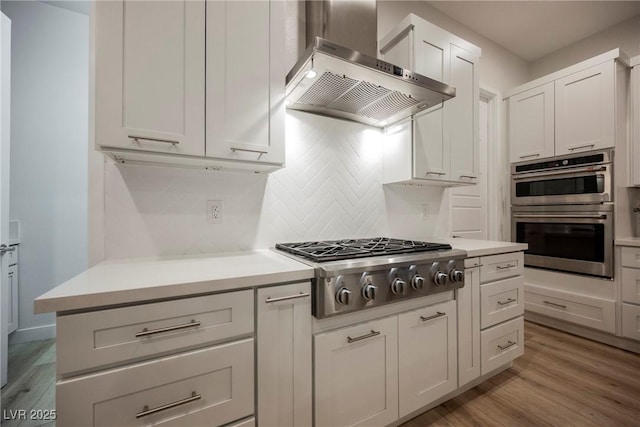 kitchen featuring white cabinetry, wall chimney range hood, stainless steel appliances, light hardwood / wood-style floors, and backsplash