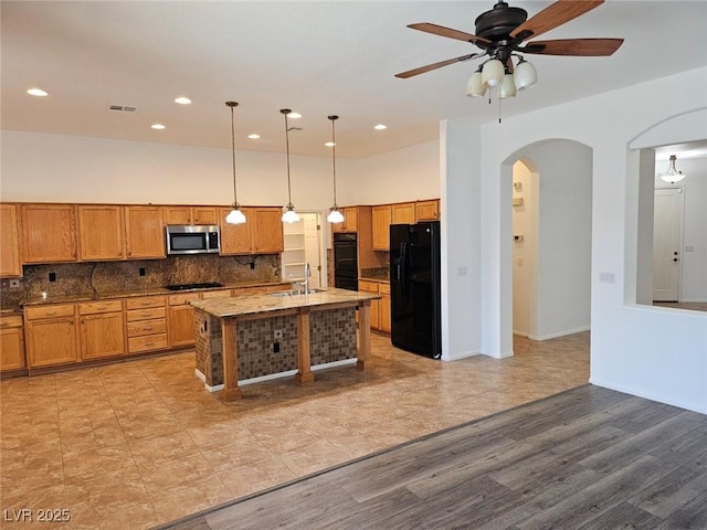 kitchen with pendant lighting, ceiling fan, backsplash, black appliances, and a center island with sink