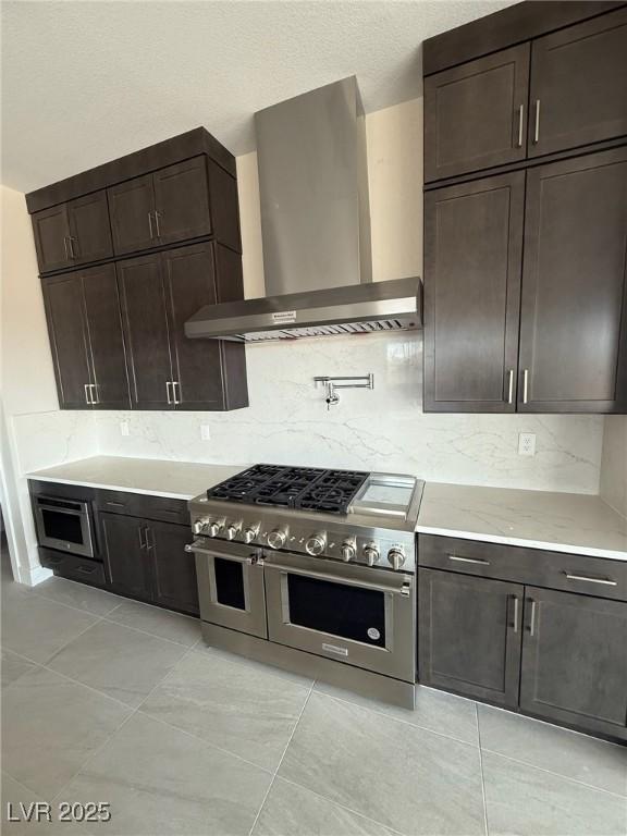 4630 Blushing Dawn Ave, Las Vegas, NV 89118 - kitchen with wall chimney range hood, range with two ovens, dark brown cabinetry, backsplash, and light tile patterned floors