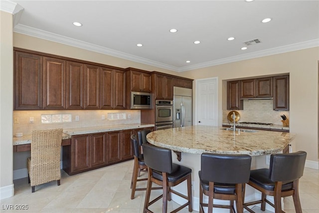 kitchen featuring an island with sink, a kitchen bar, built in appliances, crown molding, and light stone countertops