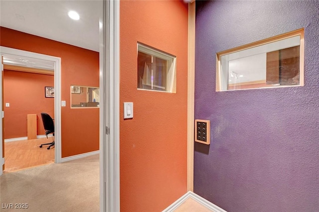 hallway featuring baseboards, a textured wall, and light colored carpet