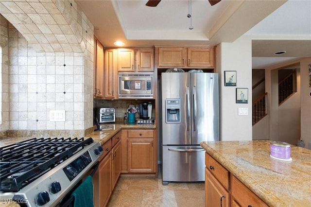 kitchen with tasteful backsplash, appliances with stainless steel finishes, a raised ceiling, ceiling fan, and light stone countertops