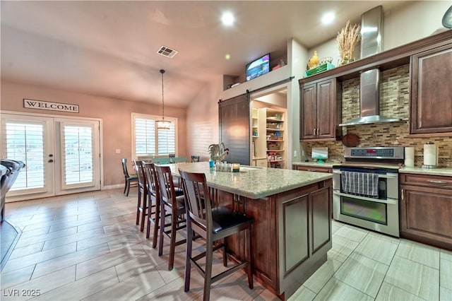 kitchen with wall chimney exhaust hood, a center island with sink, pendant lighting, a barn door, and range with two ovens