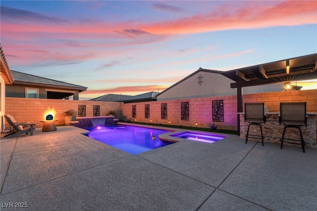 pool at dusk with a bar, a patio area, pool water feature, and an in ground hot tub