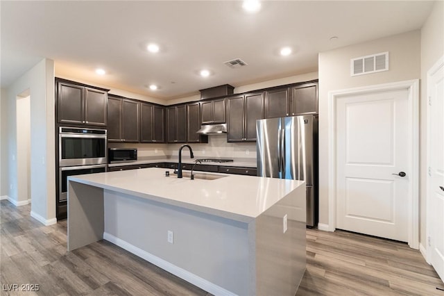 kitchen featuring appliances with stainless steel finishes, sink, a kitchen island with sink, light hardwood / wood-style floors, and dark brown cabinets