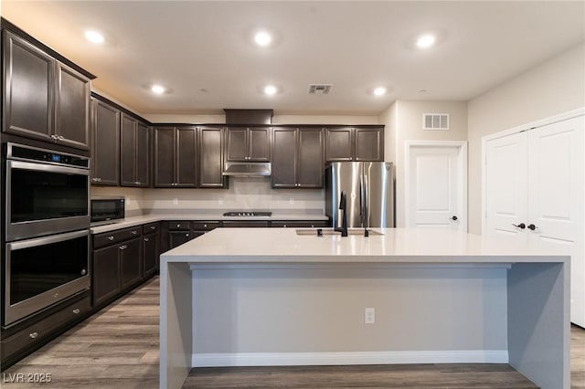 kitchen featuring stainless steel appliances, an island with sink, hardwood / wood-style flooring, and dark brown cabinetry