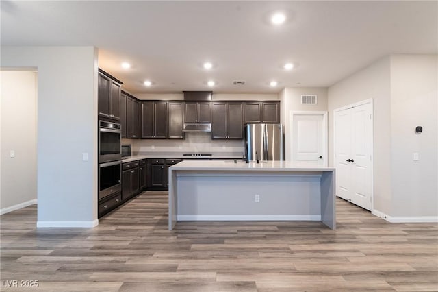 kitchen with stainless steel appliances, dark brown cabinets, a center island with sink, and light hardwood / wood-style flooring