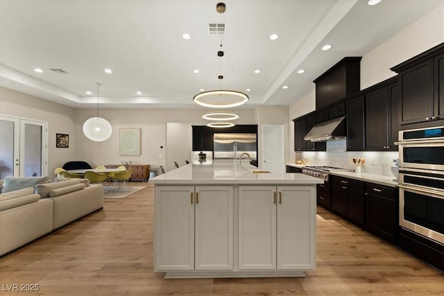 kitchen featuring pendant lighting, a tray ceiling, ventilation hood, and a center island with sink