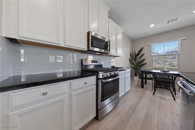 kitchen with appliances with stainless steel finishes, white cabinetry, backsplash, dark stone countertops, and light hardwood / wood-style floors