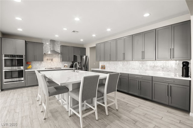 kitchen featuring a breakfast bar area, gray cabinetry, a kitchen island with sink, stainless steel appliances, and wall chimney range hood