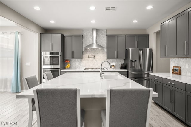 kitchen with stainless steel appliances, an island with sink, a kitchen breakfast bar, and wall chimney exhaust hood