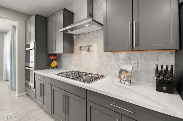 kitchen featuring wall chimney exhaust hood, gray cabinetry, appliances with stainless steel finishes, light hardwood / wood-style floors, and backsplash