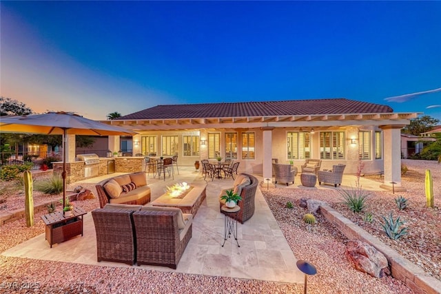 patio terrace at dusk featuring exterior kitchen, an outdoor living space with a fire pit, and grilling area