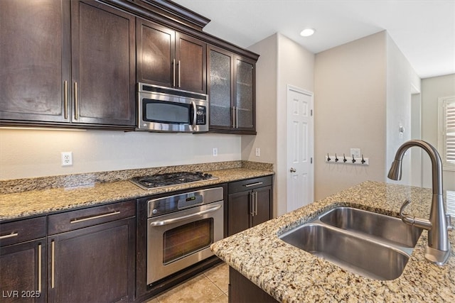 kitchen with dark brown cabinetry, light tile patterned floors, light stone counters, stainless steel appliances, and a sink