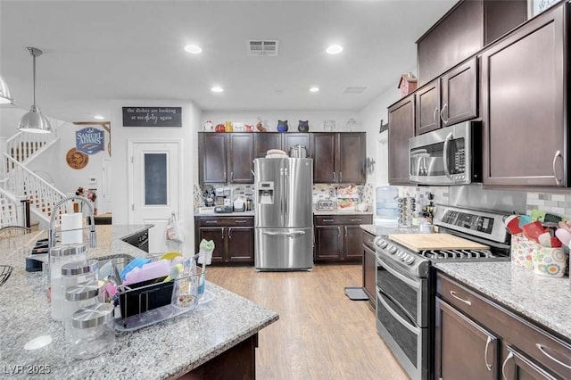 kitchen featuring appliances with stainless steel finishes, hanging light fixtures, light stone counters, dark brown cabinetry, and light hardwood / wood-style floors