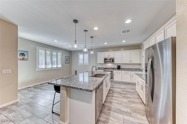 kitchen featuring pendant lighting, appliances with stainless steel finishes, a kitchen island with sink, light stone countertops, and white cabinets