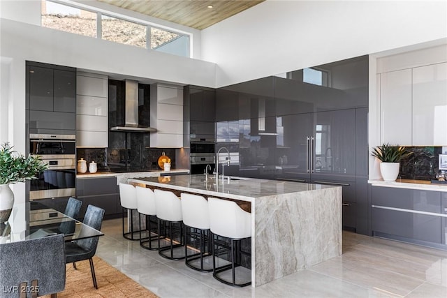 kitchen with a breakfast bar area, tasteful backsplash, an island with sink, wall chimney range hood, and a high ceiling