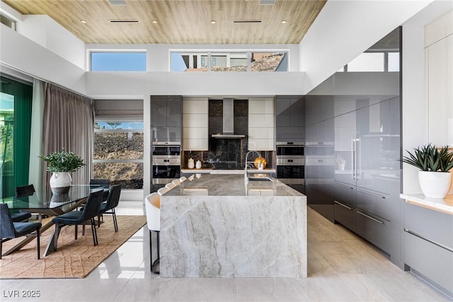 kitchen with a spacious island, sink, wooden ceiling, a towering ceiling, and wall chimney range hood