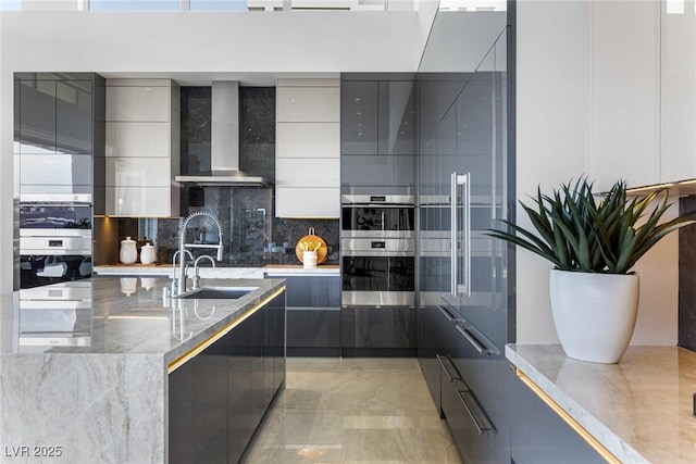 kitchen with wall chimney range hood, sink, tasteful backsplash, light stone countertops, and stainless steel double oven