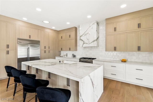 kitchen with high end appliances, tasteful backsplash, light stone counters, light wood-type flooring, and a kitchen island with sink