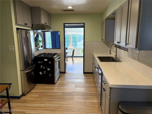 kitchen featuring stainless steel appliances, light countertops, gray cabinetry, under cabinet range hood, and a sink