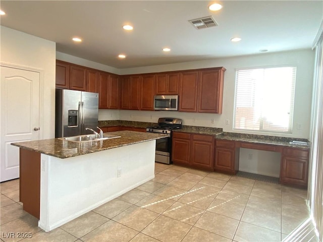 kitchen with sink, dark stone countertops, light tile patterned floors, stainless steel appliances, and a center island with sink