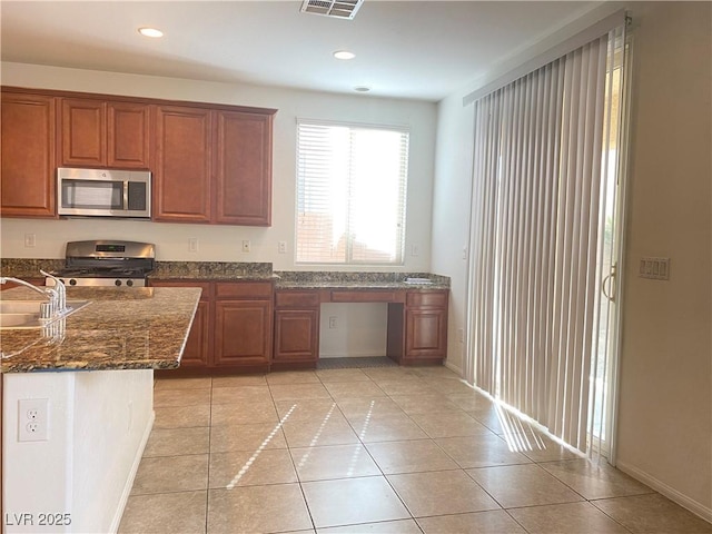 kitchen featuring sink, stainless steel appliances, built in desk, light tile patterned flooring, and dark stone counters