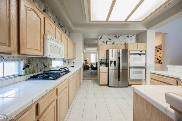 kitchen with light brown cabinetry, tile countertops, light tile patterned floors, stainless steel appliances, and backsplash