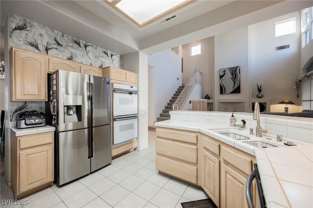 kitchen with tile counters, white double oven, stainless steel fridge, and light brown cabinets