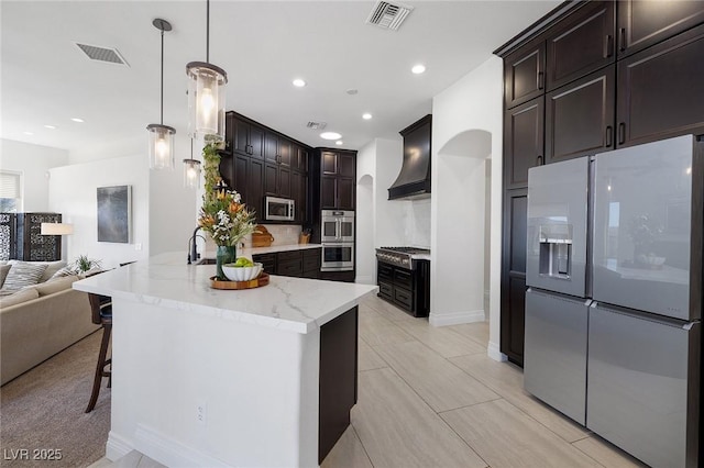 kitchen with custom exhaust hood, dark brown cabinets, hanging light fixtures, a center island with sink, and stainless steel appliances