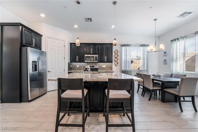 kitchen with hanging light fixtures, backsplash, stainless steel appliances, light stone countertops, and a center island with sink