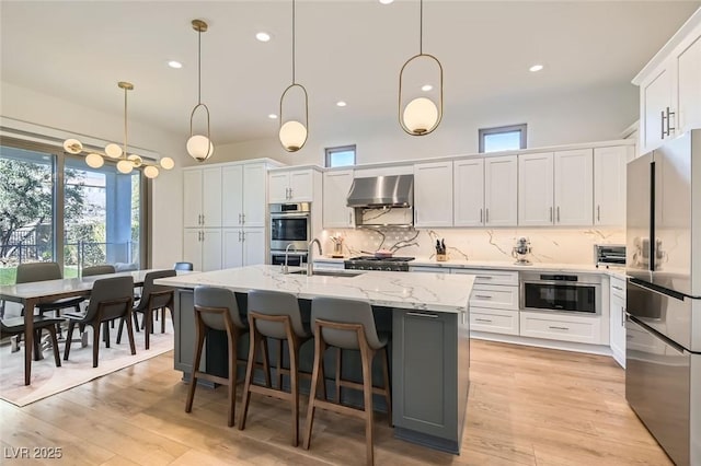 kitchen featuring wall chimney range hood, sink, a kitchen island with sink, hanging light fixtures, and stainless steel appliances