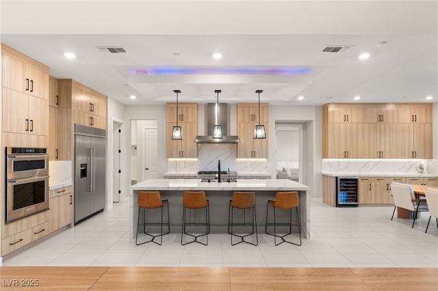 kitchen featuring a center island with sink, appliances with stainless steel finishes, light brown cabinets, and wall chimney exhaust hood