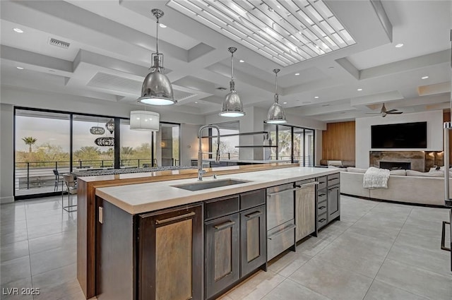 kitchen with coffered ceiling, sink, dark brown cabinets, hanging light fixtures, and a kitchen island with sink