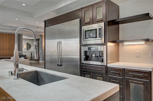 kitchen featuring built in appliances, sink, light stone countertops, and dark brown cabinetry