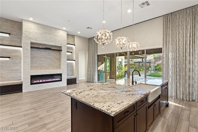 kitchen featuring dark brown cabinetry, sink, hanging light fixtures, a large fireplace, and a kitchen island with sink