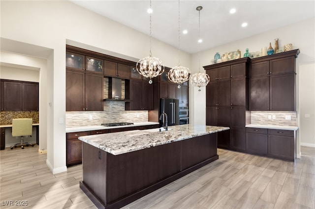 kitchen with appliances with stainless steel finishes, built in desk, an island with sink, dark brown cabinetry, and wall chimney range hood