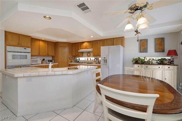 kitchen featuring white appliances, a tray ceiling, an island with sink, light tile patterned flooring, and tile countertops