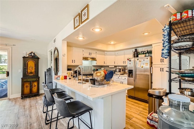 kitchen with stainless steel refrigerator with ice dispenser, under cabinet range hood, a peninsula, white cabinets, and light wood finished floors