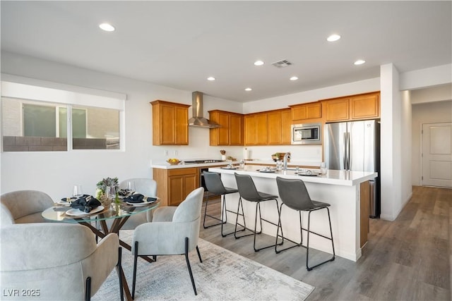 kitchen featuring wood-type flooring, a kitchen breakfast bar, a kitchen island with sink, stainless steel appliances, and wall chimney exhaust hood