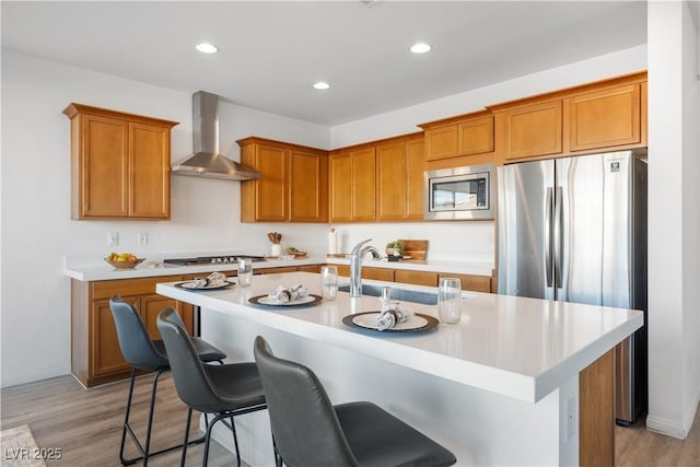 kitchen with appliances with stainless steel finishes, an island with sink, a kitchen breakfast bar, light hardwood / wood-style floors, and wall chimney exhaust hood