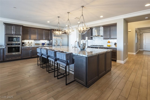 kitchen with wall chimney exhaust hood, decorative light fixtures, dark brown cabinets, a large island with sink, and appliances with stainless steel finishes