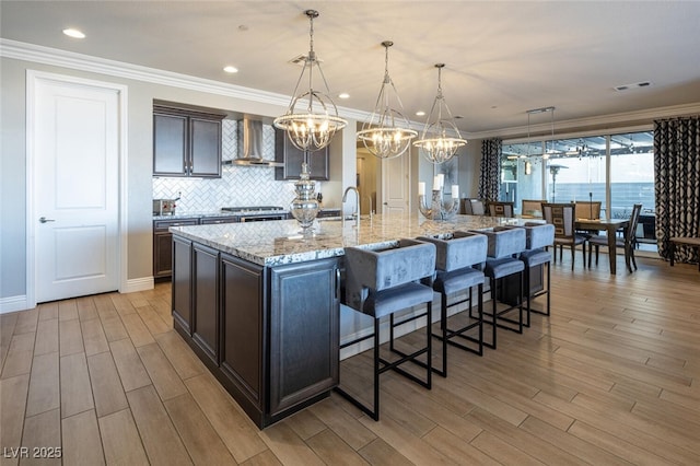 kitchen featuring decorative light fixtures, a large island, light stone counters, dark brown cabinets, and wall chimney range hood