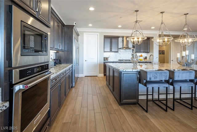 kitchen with stainless steel appliances, dark brown cabinets, wall chimney range hood, and decorative light fixtures